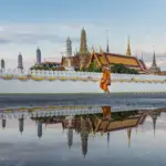 Monk walking past the gilded spires of Wat Phra Kaew in Bangkok, reflected in rainwater at dusk